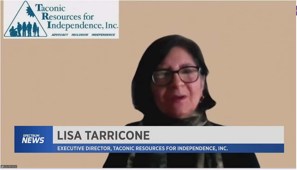 Video image of Lisa Tarricone, Executive Director of Taconic Resources for Independence, Inc. (TRI) interviewed by Spectrum News 1. Behind Lisa is the TRI logo in the upper left corner. Lisa is a white female, black hair, wearing glasses, a black sweater, and scarf.